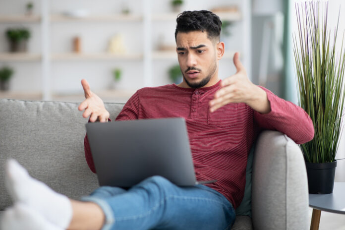 Young man throws his hands up and looks frustrated after visiting an illegal streaming service online on his laptop