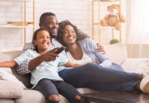 A mother, father, and young daughter smiling while sitting on the couch together, viewing streaming options to watch a new movie.
