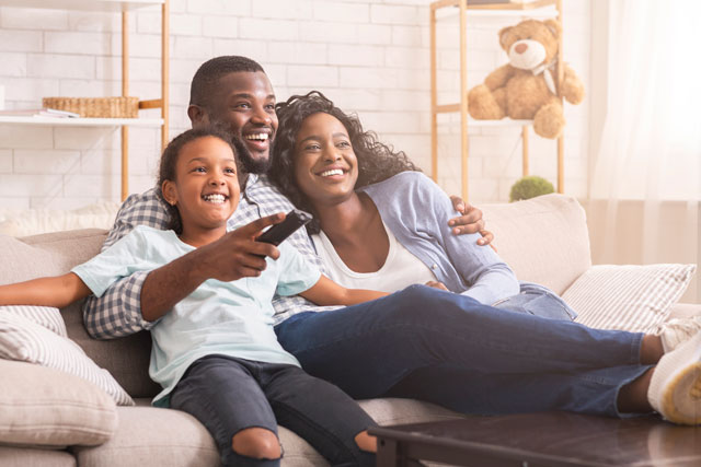 iStock-1182423333 A mother, father, and young daughter smiling while sitting on the couch together, viewing streaming options to watch a new movie.