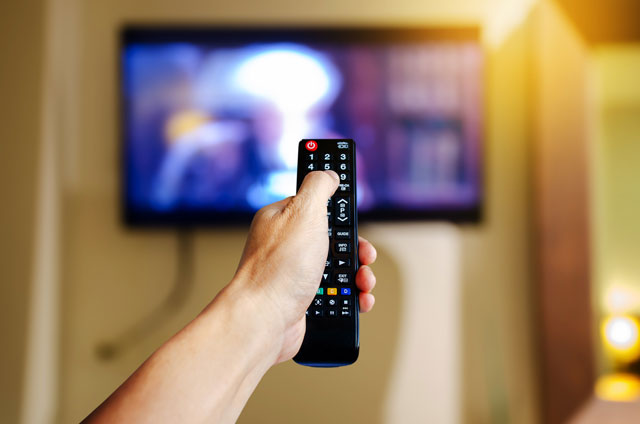A hand holding a TV remote control points at the TV to view streaming options for new shows