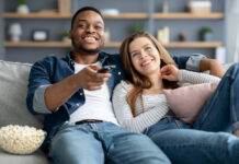 Couple sitting on couch with popcorn streaming Oppenheimer on TV