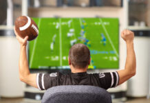 Man holding a football and wearing a football jersey cheers while watching a football game on TV