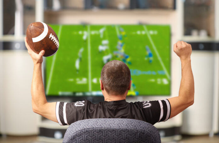 Man holding a football and wearing a football jersey cheers while watching a football game on TV