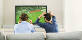 Two men sitting on a couch and reacting while watching a soccer game on TV