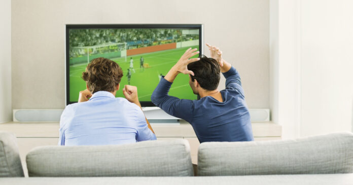 Two men sitting on a couch and reacting while watching a soccer game on TV