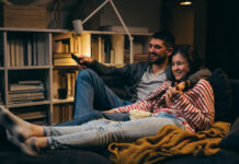 Young couple sitting together on the couch in the dark while watching a movie at home.
