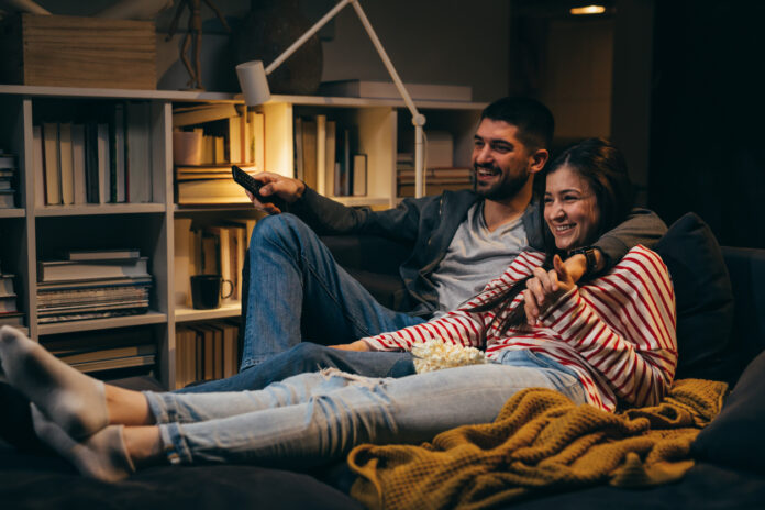 Young couple sitting together on the couch in the dark while watching a movie at home.