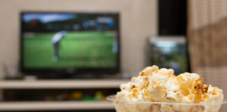 A bowl of popcorn and a TV remote with a TV broadcasting a golf tournament in the background.
