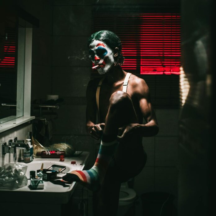 A man in clown makeup smoking a cigarette in the bathroom