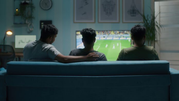 Three young men sitting on the sofa while streaming a soccer match on TV at home.