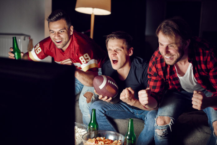 Three excited men cheer for their team while watching a football match on television.