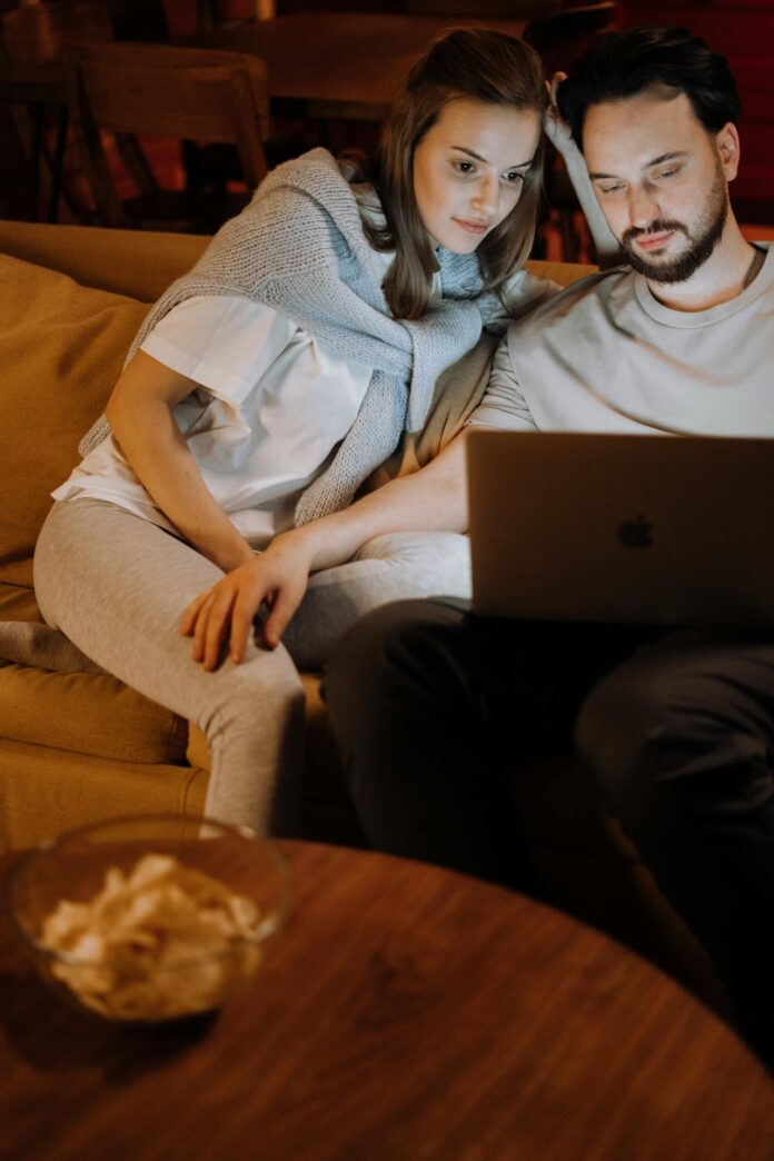 A couple sitting together at home, engaged with a laptop while watching a streaming website.