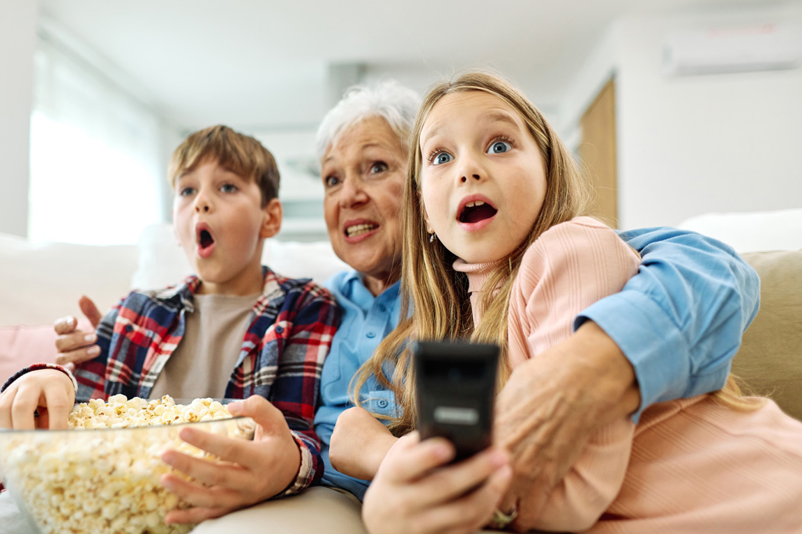 WhatIs_SafeStreaming2 Portrait of an elderly grandmother and grandchildren having fun watching tv together eating popcorn at home