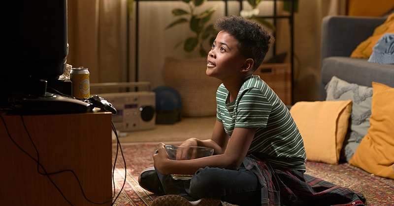 Nostalgia2 African American schoolboy sitting cross-legged on the floor watching television with a popcorn bowl in his lap in a cozy eighties living room