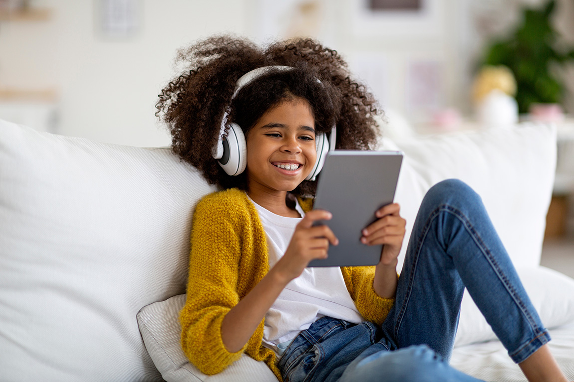 ParentalControl Happy kid school girl sitting on couch, using digital tablet and wireless headset, playing mobile game or checking newest app, home interior, copy space