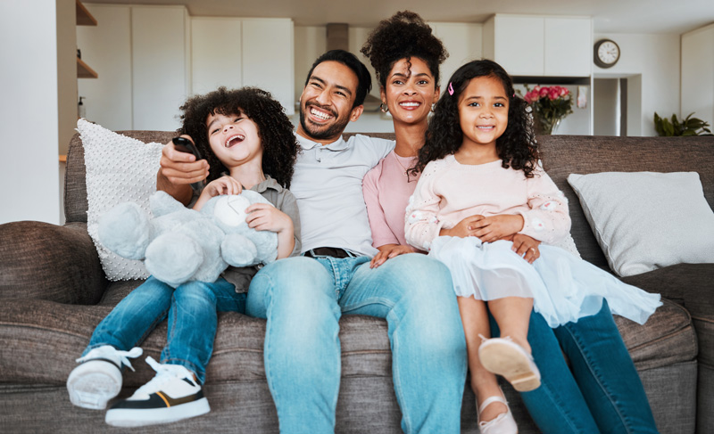 SummerCamp2 Mom, dad and children on sofa, watching tv and happy family bonding together in living room. Smile, happiness and parents relax with kids on couch, streaming television show subscription or movies.